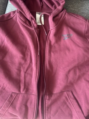 Vinröd hoodie från Reebok - Snygg vinröd hoodie från Reebok med dragkedja framtill och broderad logga på bröstet. Hoodien har huva med dragsko och två fickor framtill. Tillverkad i mjukt bomullsmaterial som är skönt att ha på sig. Perfekt för en avslappnad och sportig stil.