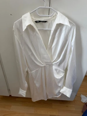 Vit omlottklänning från Zara - Snygg vit kort omlottklänning från Zara med lång ärm och djup v-ringning. Klänningen har markerad midja och klassisk krage, vilket ger en stilren och trendig look. Perfekt för dig som gillar minimalistisk stil.