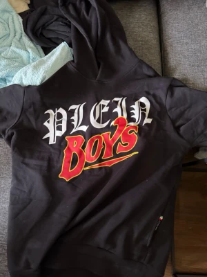 Svart Plein Boys hoodie med tryck - Svart hoodie från Plein med stor vit och röd text 'PLEIN BOYS' på bröstet. Hoodien har huva och en liten Plein-logga vid fickan med italienska flaggans färger. Perfekt för dig som gillar streetwear och vill sticka ut. Nypris 9000