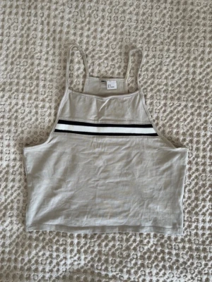 Beige linnetopp med rand - Snygg beige linnetopp från H&M med smala axelband och en svartvit rand över bröstet. Toppen är croppad och har en sportig vibe, perfekt till jeans eller shorts. Tillverkad i mjuk bomull för en skön känsla.