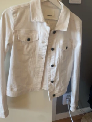 Vit jeansjacka från object  - Snygg vit jeansjacka från Bik Bok med klassisk krage, svarta knappar och två bröstfickor med lock. Jackan har raka ärmar och en figurnära passform som ger en clean och fräsch look. Perfekt att slänga över en t-shirt eller hoodie.