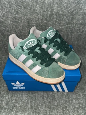 Adidas Campus gröna i mocka - Snygga gröna Adidas Campus  i mocka med vita klassiska ränder på sidorna och mörkgröna skosnören. Skorna har en beige gummisula och Adidas logga på plösen. Perfekta för dig som gillar retrostil och streetwear.