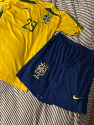 Brasilien fotbollströja & shorts  - Säljer ett Brasilien fotbollskit med gul tröja och blå shorts. Tröjan har nummer 23 och namnet Kaká på ryggen, samt gröna detaljer och Brasilien-emblem. Shortsen är blå med gul och Brasilien-märke. Perfekt för fotbollsträning eller match!
