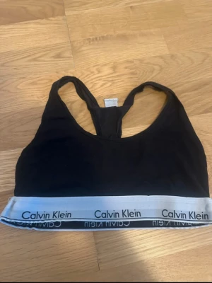 Svart sport-bh från Calvin Klein - Svart sport-bh från Calvin Klein med klassisk brottarrygg och bred resår med logotyp i vitt och svart. Mjuk och bekväm modell, perfekt för en aktiv vardag. Tillverkad i bomullsblandning för extra komfort.