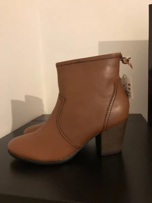 Bruna stövletter från 5th Avenue - Snygga bruna stövletter i äkta läder från 5th Avenue. Modellen har rund tå, dragkedja bak och markerade sömmar. Klack i trälook ger extra höjd och stil. Perfekta för dig som vill ha en klassisk och trendig look.