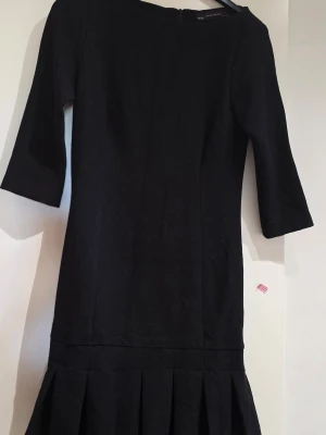 Svart klänning med plisserad nederdel - Stilren svart klänning med trekvartsärm och plisserad nederdel. Klänningen har en rak passform och är knälång, perfekt för en trendig look. Tillverkad i ett mjukt tyg som ger en snygg siluett.