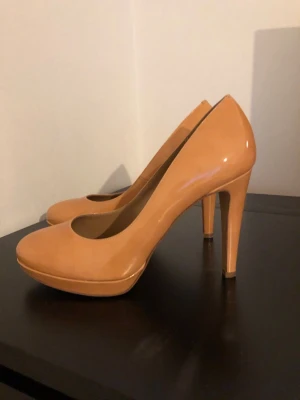 Beiga pumps från a Pair - Snygga pumps i beige lack från a Pair med rund tå och hög klack. Skorna har en klassisk siluett och platåsula framtill som ger extra höjd. Perfekta för dig som gillar stilrena och eleganta skor med en modern touch.