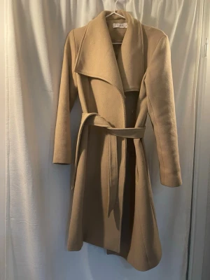 Beige kappa från Ellos med skärp - Stilren beige kappa från Ellos i storlek 34. Kappan har ett brett slag, knytbälte i midjan och är helfodrad. Tillverkad i ett mjukt material som ger en klassisk och elegant look. Perfekt för kyligare dagar och enkel att matcha med olika outfits.