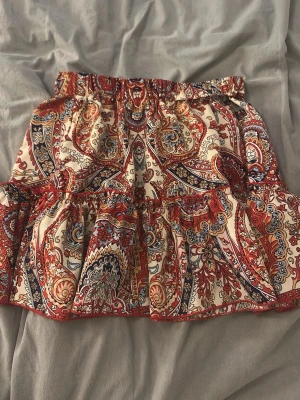 Mönstrad kortkjol med volang - Kortkjol med färgstarkt paisleymönster i rött, blått och beige. Kjolen har volang nertill och resår i midjan för en bekväm passform. Perfekt för dig som gillar boho-stil och vill sticka ut med snygga detaljer.