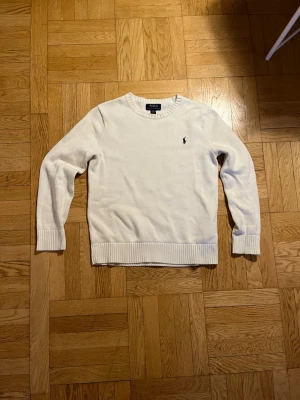 Vit stickad tröja från Polo Ralph Lauren - Klassisk vit stickad tröja från Polo Ralph Lauren med rund halsringning och ribbade muddar. Tröjan har den ikoniska mörkblå logotypen broderad på bröstet och är tillverkad i mjukt material. Perfekt för en stilren och enkel look. Passar dig perfekt om du är ungefär 164cm