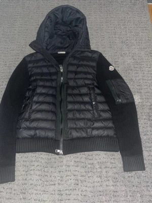 Svart pufferjacka från Moncler - Säljer en svart pufferjacka från Moncler med stickade ärmar och huva. Jackan har quiltad front, två fickor med dragkedja och en extra ficka på ärmen med Moncler-logga. Materialet är en mix av nylon och stickad ull, perfekt för kalla dagar.