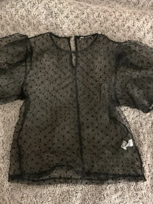 Svart transparent blus från Zara - Snygg svart  blus från Zara med puffiga korta ärmar och små prickar i tyget. Blusen har knäppning bak i nacken. Perfekt att styla lager på lager eller som den är