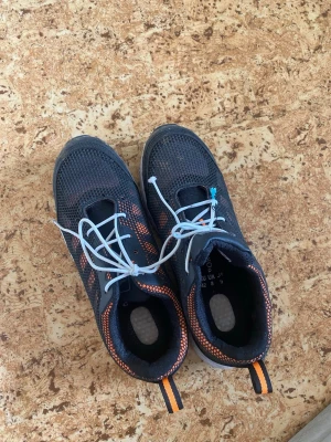 Sneaker for work bulding - Säljer ett par svarta sneakers med orange meshdetaljer och vita snören. Skorna har en vit sula och dragflik bak med orange och svart rand. Ovandelen är i ventilerande textilmaterial och modellen är lågskuren för en sportig look.