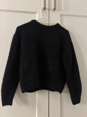 Svart fluffig stickad tröja - Mysig svart stickad tröja med fluffig textur och rund hals. Tröjan har långa ärmar och ribbade muddar vid ärmslut och nederkant. Perfekt för kyliga dagar och enkel att matcha med olika outfits.