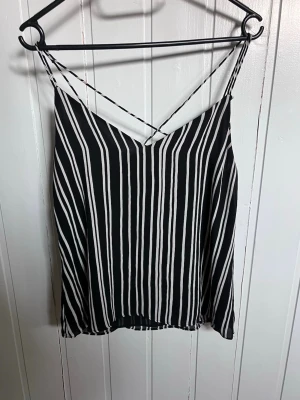 svartvit linnetopp med smala band - Säljer en svart och vit randig linnetopp med tunna axelband som korsas i ryggen. Toppen har en lös passform och är perfekt för varma dagar. Materialet känns lätt och luftigt, och toppen har en snygg v-ringning framtill.