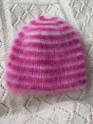 Rosa och vit randig fluffig mössa - Mysig virkad mössa i fluffigt garn med rosa och vita ränder. Mössan har en klassisk form och är perfekt för att hålla värmen under kalla dagar. Materialet är i alpacka för extra mjuk känsla.
