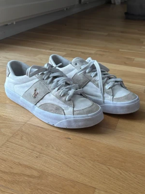Beige sneakers från Polo Ralph Lauren - Snygga beige sneakers från Polo Ralph Lauren med klassisk låg profil. Skorna har mockadetaljer, vita partier i textil och den ikoniska loggan broderad på sidan. Tjock vit sula och matchande beigea skosnören ger en clean och stilren look.