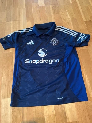 Manchester United bortatröja Garnacho - Snygg mörkblå Manchester United bortatröja från Adidas med Garnacho och nummer 17 på ryggen. Tröjan har vita detaljer, klubbmärke på bröstet, Snapdragon sponsortryck och klassiska Adidas-ränder på axlarna. Tillverkad i lätt och ventilerande polyester.