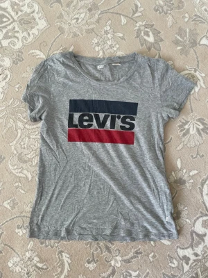 Grå Levi's t-shirt med logga - Snygg grå t-shirt från Levi's med klassisk stor logga i blått, vitt och rött framtill. T-shirten har rund halsringning och korta ärmar. Tillverkad i mjuk bomull som känns skön mot huden. Perfekt till jeans eller shorts.