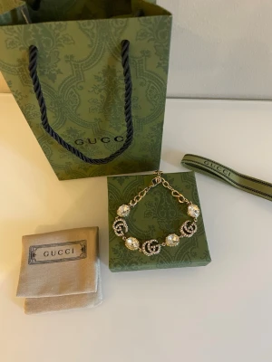 Gucci armband med pärlor och kristaller - Lyxigt armband från Gucci i guldfärgad metall med stora glittrande pärlor och ikoniska GG-loggor täckta av små kristaller. Armbandet har en chunky kedja och detaljer som verkligen sticker ut. Perfekt för dig som vill ha något extra till din stil.