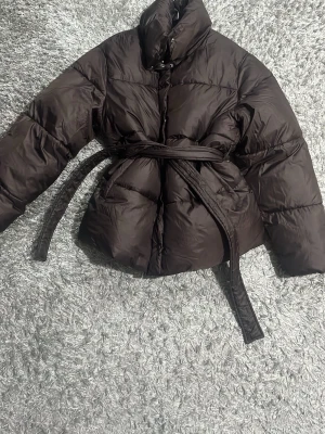 Brun pufferjacka med bälte - Snygg brun pufferjacka med hög krage och tillhörande bälte i midjan. Jackan har en quiltad design och stängs med knappar framtill. Perfekt för kalla dagar och ger en trendig siluett.