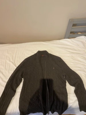 Mörkbrun kabelstickad Morris kofta - Snygg mörkbrun kabelstickad kofta från Morris med dragkedja framtill och diskret logga på bröstet. Koftan har lång ärm och ribbade muddar vid ärmslut och nederkant. Perfekt för lager på lager och en klassisk look. Äkta Morris! Säljs för den inte används så mycket