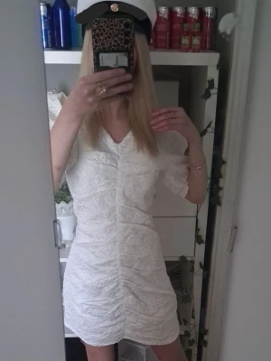 Vit kort spetsklänning - Säljer en vit kort klänning med v-ringning och fina spetsdetaljer. Klänningen har korta puffärmar och en figurnära passform som framhäver siluetten. Perfekt till student eller sommarens alla tillfällen.