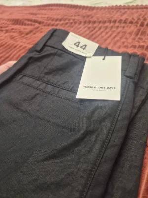 These Glory Days T Faifox svarta byxor, storlek 44 - Snygga mörkgrå chinos från These Glory Days med klassisk rak passform och bälteshällor. Byxorna har två sidofickor och en bakficka, perfekta för en clean och stilren look. Materialet känns mjukt och slitstarkt, vilket gör dem riktigt bekväma.