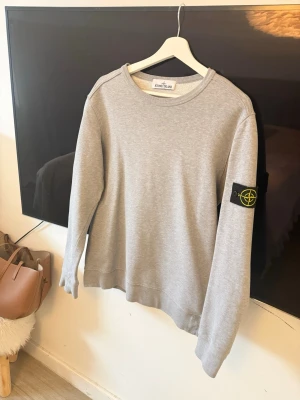 Grå sweatshirt från Stone Island - Säljer en stilren grå sweatshirt från Stone Island med klassisk rund hals och lång ärm. Tröjan har den ikoniska svarta patchen med gult Stone Island-märke på vänster ärm. Materialet är mjukt och bekvämt, perfekt för en avslappnad look.