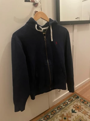 Mörkblå zip-hoodie från Polo Ralph Lauren - Mörkblå hoodie med dragkedja från Polo Ralph Lauren. Klassisk design med huva och vita snören, samt fickor framtill. Liten röd logga broderad på bröstet. Perfekt för en avslappnad och stilren look.