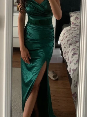 Grön långklänning med slits - Elegant grön långklänning i glansigt material med omlottdetalj upptill och markerad midja. Klänningen har en hög slits framtill och smickrande drapering. Min balklänning, använd en gång.