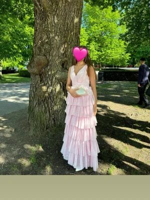 Ljusrosa volangklänning med djup rygg - Säljer en lång, ljusrosa klänning med flera lager volanger och djup v-ringning fram samt öppen rygg. Klänningen är från bubbleroom och heter ”Tulle Frill Gown” i färgen ljusrosa, köpt förra året. Klänningen har breda axelband och är tillverkad i ett tunt, luftigt material som ger en romantisk look. Perfekt för speciella tillfällen så som exempelvis bal eller bröllop. Jag själv hade denna på balen år 2025 🩷.                               Nypris: 1799kr