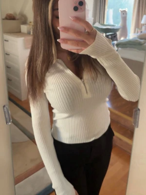 Vit  tröja med dragkedja - Säljer en vit, ribbad långärmad tröja med dragkedja framtill. Tröjan har figurnära passform och mjukt stretchigt material som sitter snyggt på kroppen. Perfekt till jeans för en clean och stilren look.