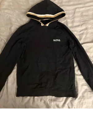 Svart hoodie från BOSS med beige kant - Snygg svart hoodie från BOSS med beige och vit rand längs huvan. Klassisk känguruficka framtill och BOSS-logga broderad på bröstet. Perfekt för en avslappnad och stilren look. Tillverkad i mjukt bomullsmaterial.