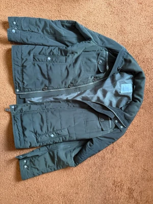 Svart pufferjacka från J.Lindeberg - Snygg svart pufferjacka från J.Lindeberg med flera fickor framtill, dragkedja och knappar. Jackan har en hög krage och en patch med logga på ärmen.