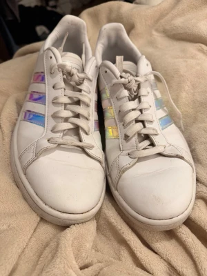 Vita Adidas sneakers med holografiska ränder - Säljer ett par vita Adidas sneakers med klassiska tre holografiska ränder på sidorna som skiftar i blått, lila och gult. Skorna har rund tå, vita snören och är tillverkade i slätt lädermaterial. Perfekta för dig som vill sticka ut lite extra.använda en gång på studenten. 