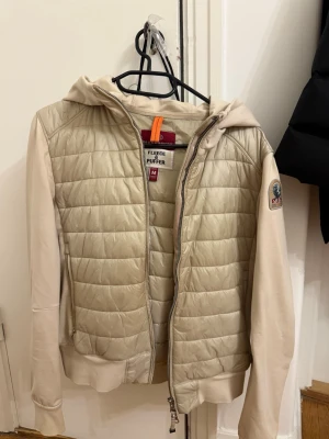 Beige pufferjacka med huva från Parajumpers - Snygg beige pufferjacka från Parajumpers med quiltad front och rygg, mjuka ärmar och huva i sweatshirtmaterial. Jackan har dragkedja framtill, ribbade muddar och en patch med logga på ärmen. Perfekt för kalla vinterdagar och riktigt skön att bära.