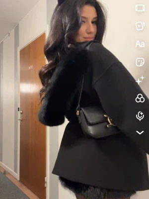 Svart kappa med fuskpälsdetaljer - Stilren svart kappa med markerad midja och knytskärp. Kappan har breda ärmar med fuskpälsdetaljer vid axlarna och en mjuk, något oversized passform. Perfekt för dig som vill ha en trendig och elegant look. Två stora fickor framtill. Köpt för 2000kr, pris kan diskuteras