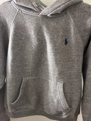 Grå hoodie från Polo Ralph Lauren - Klassisk grå hoodie från Polo Ralph Lauren med mörkblå logga broderad på bröstet. Hoodien har en stor magficka och ribbade muddar vid ärmslut och nederkant. Tillverkad i mjuk bomullsblandning för maximal komfort.