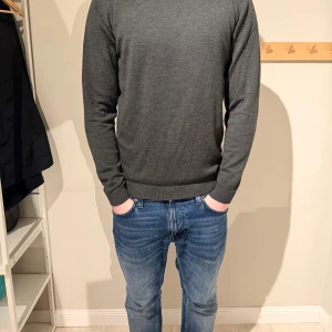 Mörkgrå/grön stickad tröja från A Days March  - En stilren mörkgrå/grön stickad tröja med rund halsringning och ribbade muddar vid ärmslut och nederkant. Tröjan har en klassisk passform och är perfekt för lager på lager. Materialet ser ut att vara mjukt och bekvämt, troligen bomull eller ullblandning. Tröjan är i storlek L och har inga skavanker utom ett litet hål vid ryggen men går att fix hemma 