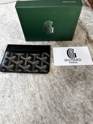 Svart plånbok  - Snyggt svart plånbok med det klassiska mönstret i vitt och beige. Tillverkat i exklusivt läder och canvas, perfekt för kort och ID. levereras med originalask och certifikat.