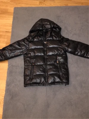 Svart dunjacka med huva - Säljer en svart dunjacka med huva och dragkedja framtill. Jackan har två sidofickor med dragkedja och quiltad design som ger en puffig look. Perfekt för kalla vinterdagar och riktigt skön att bära. Köpt från nätet,