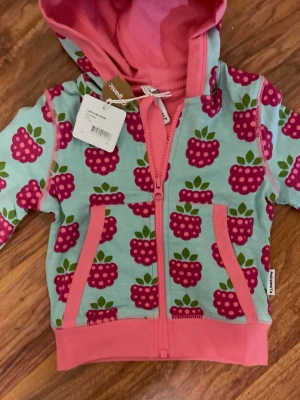 Mönstrad hoodie från Maxomorra - Färgglad hoodie från Maxomorra med hallonmönster i rosa och grönt på ljusblå botten. Tröjan har rosa muddar, dragkedja framtill, fickor och en mysig huva med rosa insida. Perfekt för dig som gillar lekfull stil och sköna material.