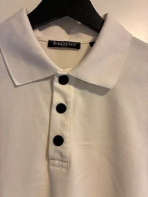 Vit pikétröja från Balmain - Vit pikétröja från Balmain med svarta knappar framtill och svart logotyptext på ärmsluten. Klassisk krage och korta ärmar. Tillverkad i bomull för en bekväm känsla och stilren look.