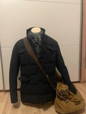 Massimo dutti jacka - Stilren svart pufferjacka med flera stora fickor framtill och hög krage. Jackan har en quiltad design och stängs med knappar. Perfekt för kalla dagar och ger en modern look. Materialet ser ut att vara syntet med vaddering för extra värme.