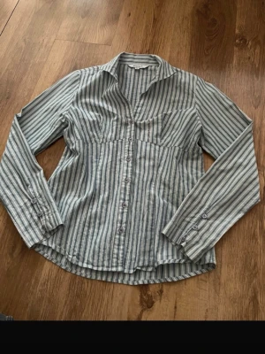 Randig skjorta med knappar - Snygg skjorta med smalrandigt mönster i blått, vitt och grått. Skjortan har lång ärm, klassisk krage och knäppning framtill samt vid ärmslut. Tillverkad i ett mjukt bomullstyg som känns skönt mot huden. Perfekt för en avslappnad men ändå stilren look.