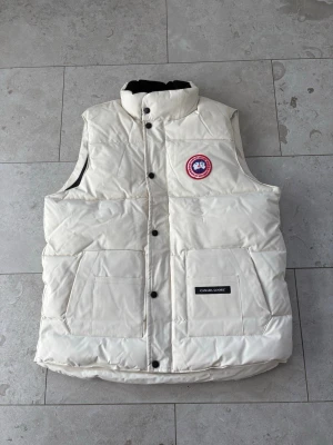 Vit dunväst från Canada Goose - Snygg vit dunväst från Canada Goose med hög krage och tryckknappar framtill. Västen har två stora framfickor, klassisk logopatch på bröstet och quiltad design. Perfekt för lager-på-lager och håller dig varm under kyliga dagar.