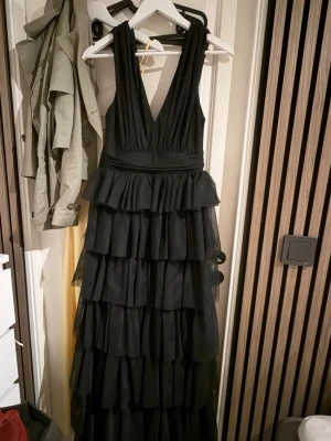 Svart lång balklänning från Bubbleroom - Säljer en svart långklänning från Bubbleroom med djup v-ringning och flera lager volanger. Klänningen har ärmlös design och markerad midja, perfekt för speciella tillfällen. Materialet känns mjukt och faller snyggt.