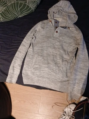 Ljusgrå stickad hoodie med knappar - Säljer en ljusgrå stickad hoodie med knappslå och bruna detaljer vid halsen. Hoodien har magficka framtill, ribbade muddar och en skön, avslappnad passform. Perfekt för dig som gillar en enkel men snygg stil.