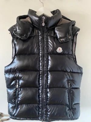 Moncler Väst - Snygg svart dunväst från Moncler med glansig finish, hög krage och avtagbar huva. Västen har två dragkedjefickor fram och klassisk Moncler-logga på bröstet. Skriv för mer info